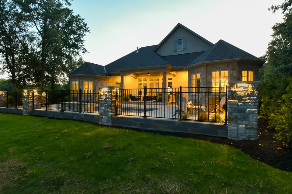 Evening view of poolside landscape with lit house, fence, stone piers, pool, and patio